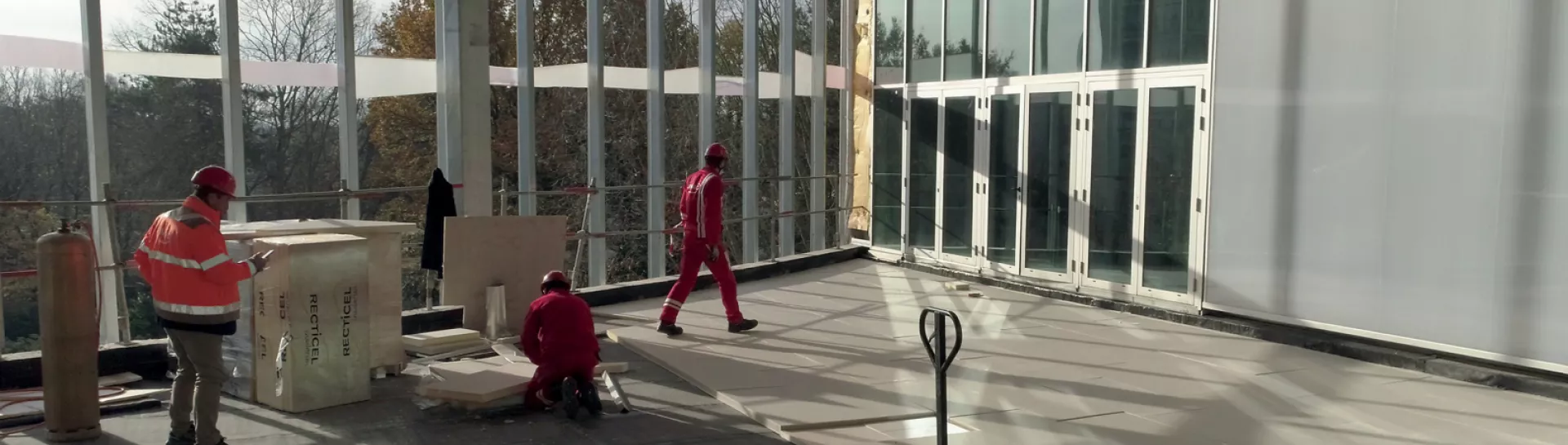 Workers in safety gear fixing flooring in a large, sunlit room with glass walls.