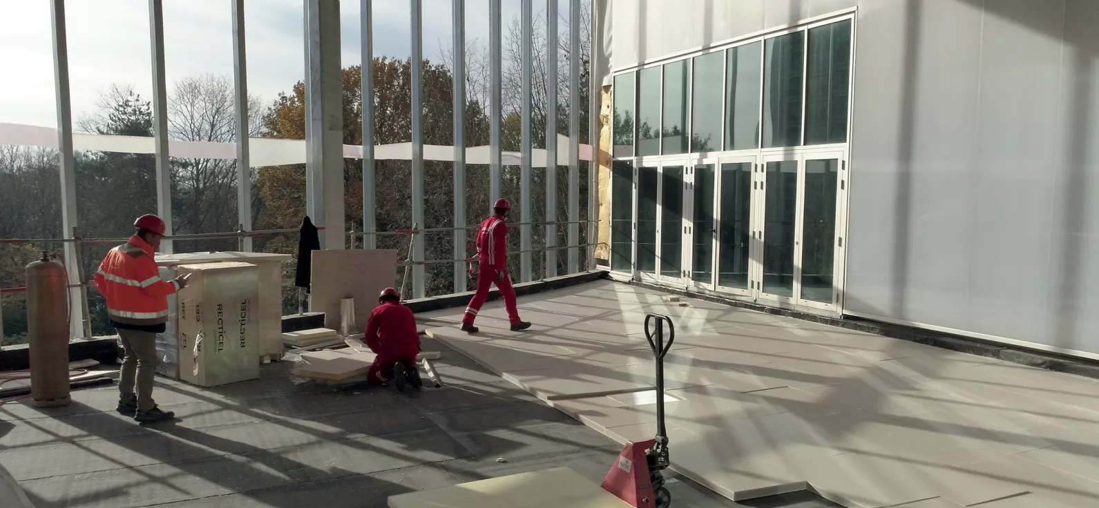 Workers in safety gear fixing flooring in a large, sunlit room with glass walls.
